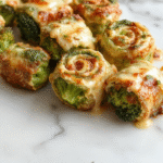 Colorful broccoli cheese pinwheels on a white plate, featuring golden-brown flaky pastry filled with vibrant green broccoli and melted cheese, styled with a sprinkle of fresh herbs for a tempting presentation.